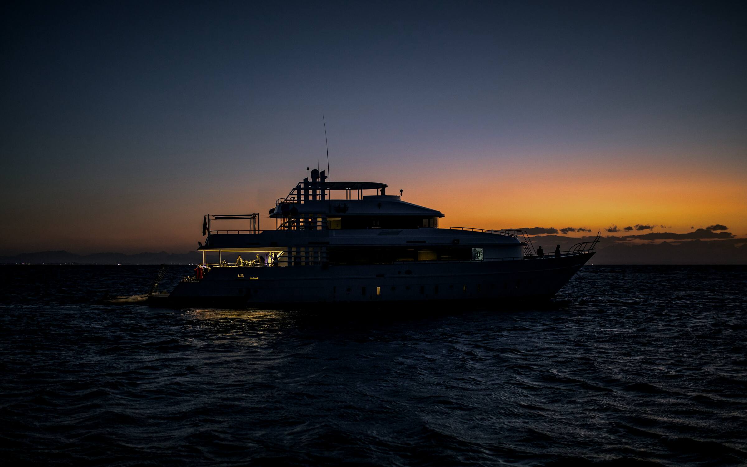 A motor yacht at anchor on open water at dusk, its aft deck lit warm gold against the last orange light on the horizon.